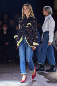 Female model walking a runway in a dark embroidered blazer, cuffed jeans, and red high heels.