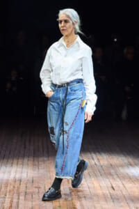 Older woman walking a runway in a white button-down shirt and embellished blue jeans during a fashion show.