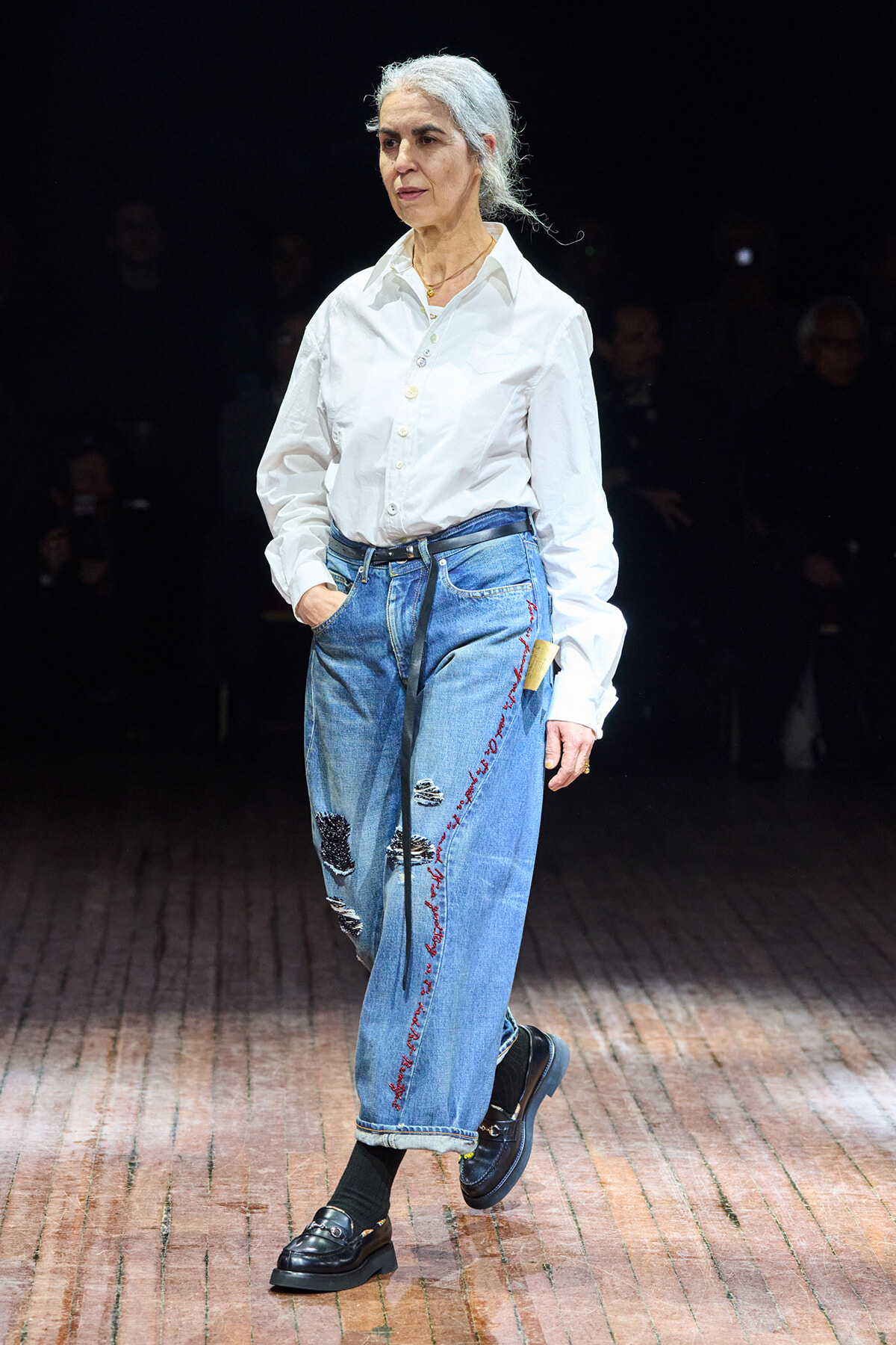 Older woman walking a runway in a white button-down shirt and embellished blue jeans during a fashion show.