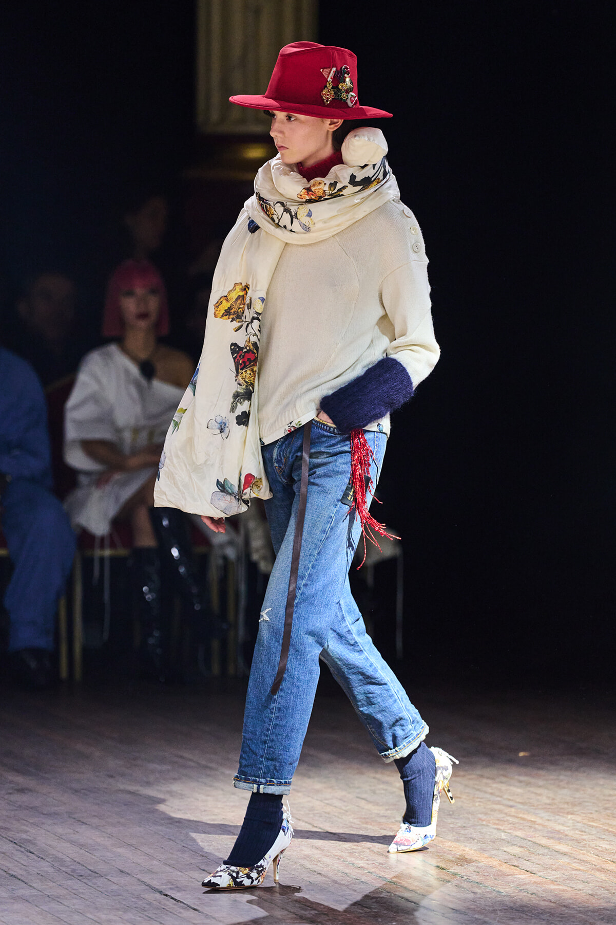 Model walks a fashion show runway in a red wide-brim hat, cream knit sweater, floral scarf, rolled jeans, and floral heels.