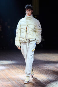 Model walks the runway in an oversized cream puffer jacket, floral hem, and floral-heeled shoes with a spiky crown accessory.