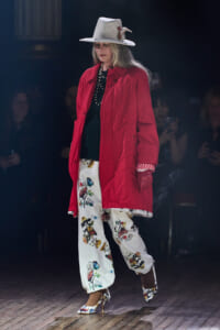 Model walking the runway in a red coat, white hat with a feather, and bird-printed pants with floral heels.