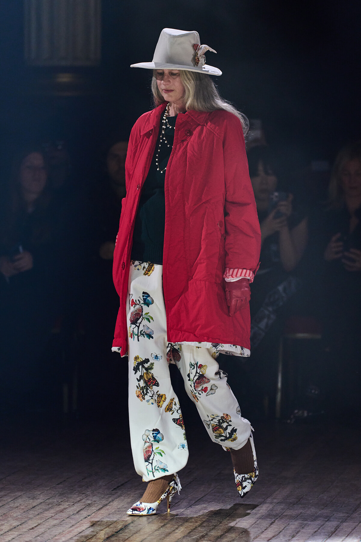 Model walking the runway in a red coat, white hat with a feather, and bird-printed pants with floral heels.