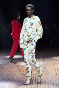 Model walks the runway in a white floral print sweatsuit with butterfly designs, accessorized with a spiked gold crown and chunky jewelry.