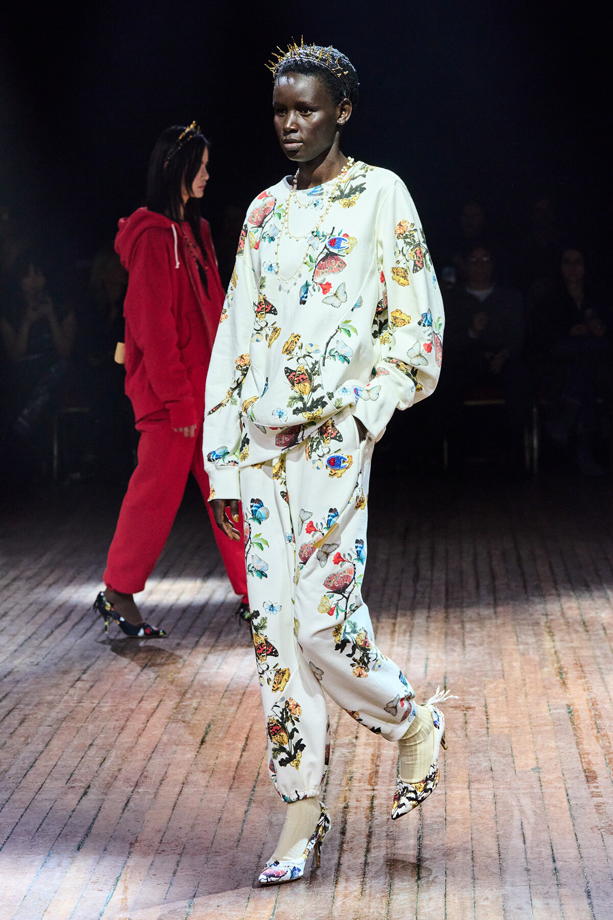 Model walks the runway in a white floral print sweatsuit with butterfly designs, accessorized with a spiked gold crown and chunky jewelry.