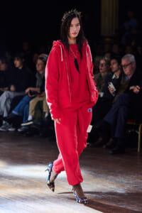 Model walking a runway in a red velour tracksuit with a crown headpiece and floral heels on the catwalk.