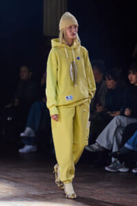 Model walking a fashion runway in a yellow hoodie and matching sweatpants, beige knit beanie, and long pendant necklace.
