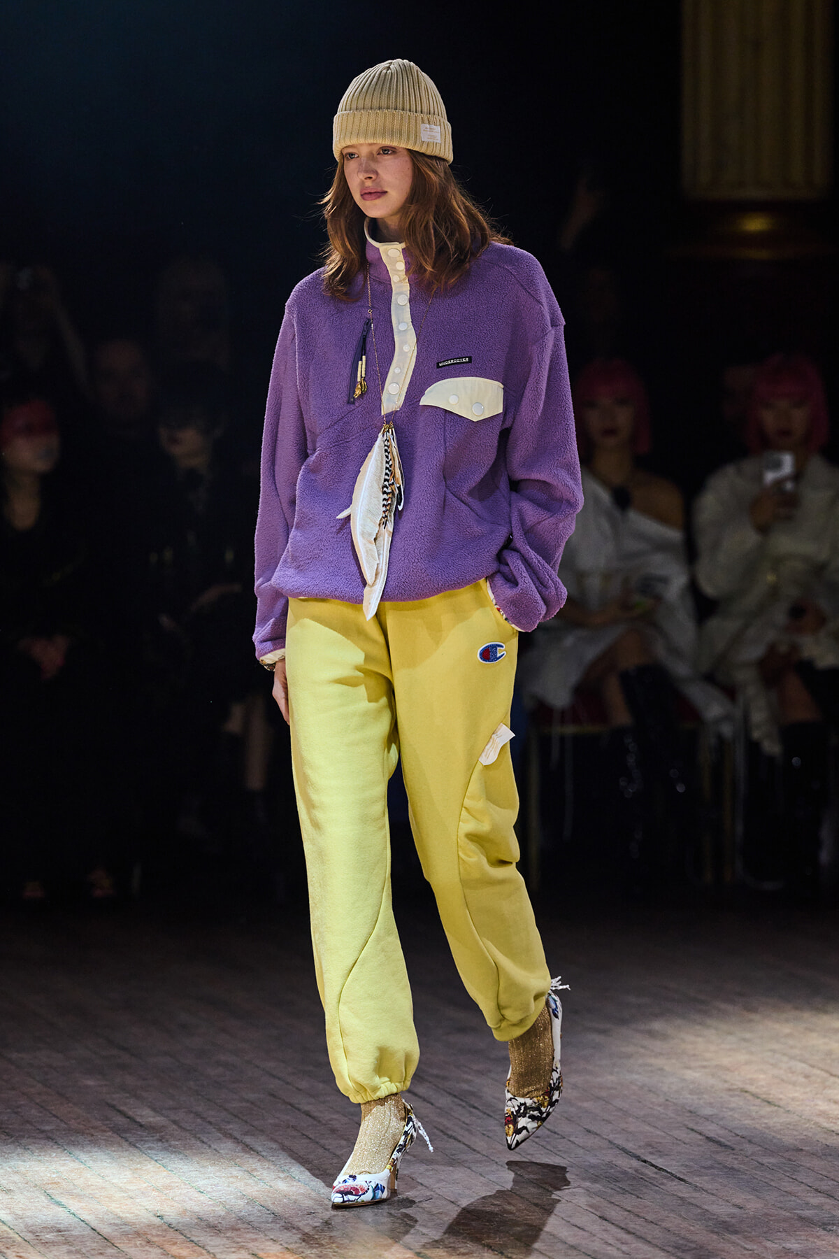 Model walking the runway in a purple fleece pullover, yellow sweatpants, beige beanie, and printed heeled shoes on a wooden catwalk.