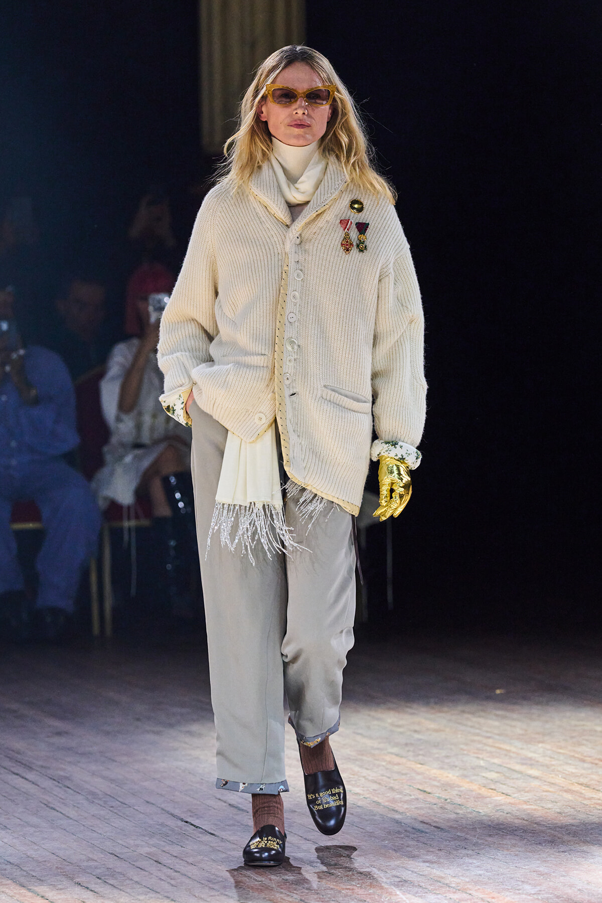 Model walks the runway in a cream oversized knit cardigan with medals, turtleneck, and light gray trousers; orange sunglasses and gold gloves stand out at the sides.
