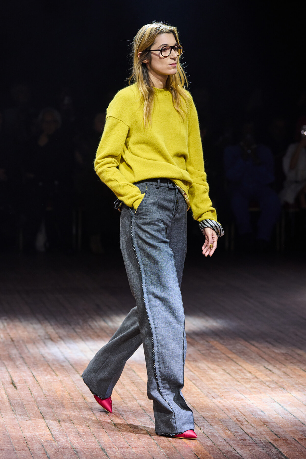 Model walks a fashion show runway in a bright yellow oversized sweater, wide-leg gray jeans with side seams, pink pointed-toe heels, and dark-framed glasses.