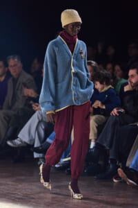 Model walks runway wearing a chunky blue cardigan with cream trim, burgundy pants with fringe, snake-print heels, and a beige knit cap.