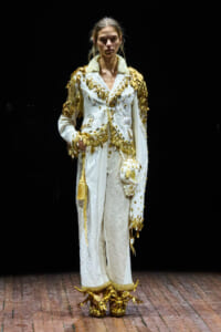 Model in a white, heavily embroidered suit with gold appliqués and shoulder embellishments on a runway.