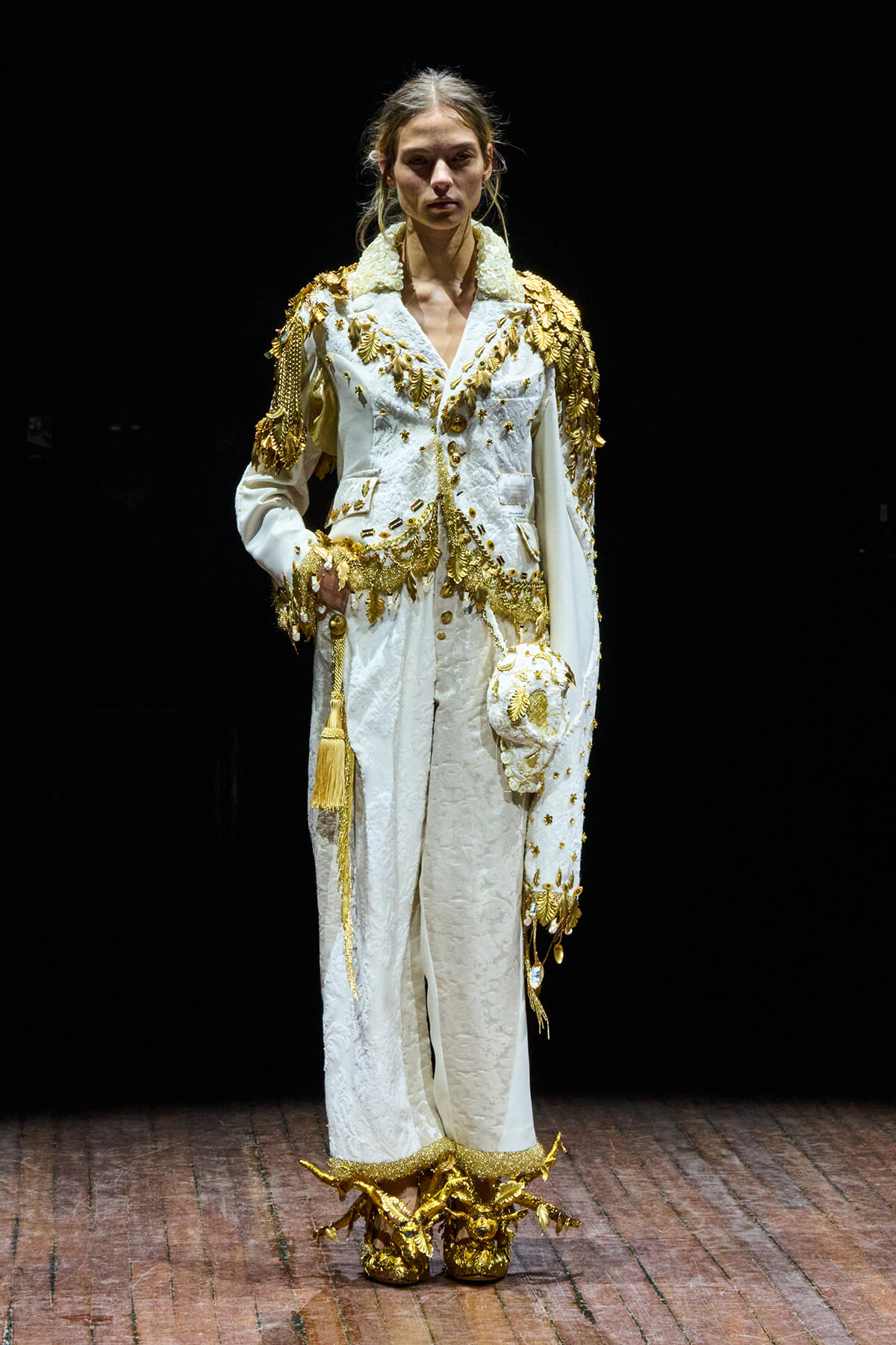 Model in a white, heavily embroidered suit with gold appliqués and shoulder embellishments on a runway.