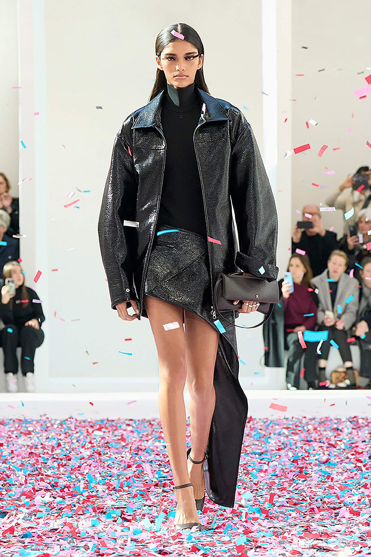Model on a runway wearing a black turtleneck, oversized shiny black jacket, and asymmetrical skirt; colorful confetti falls around her.