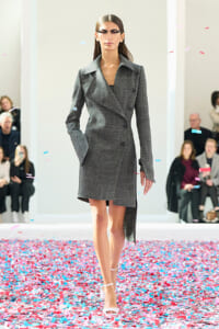 Model walks a fashion runway in a gray double-breasted coat; pink and blue confetti covers the floor.