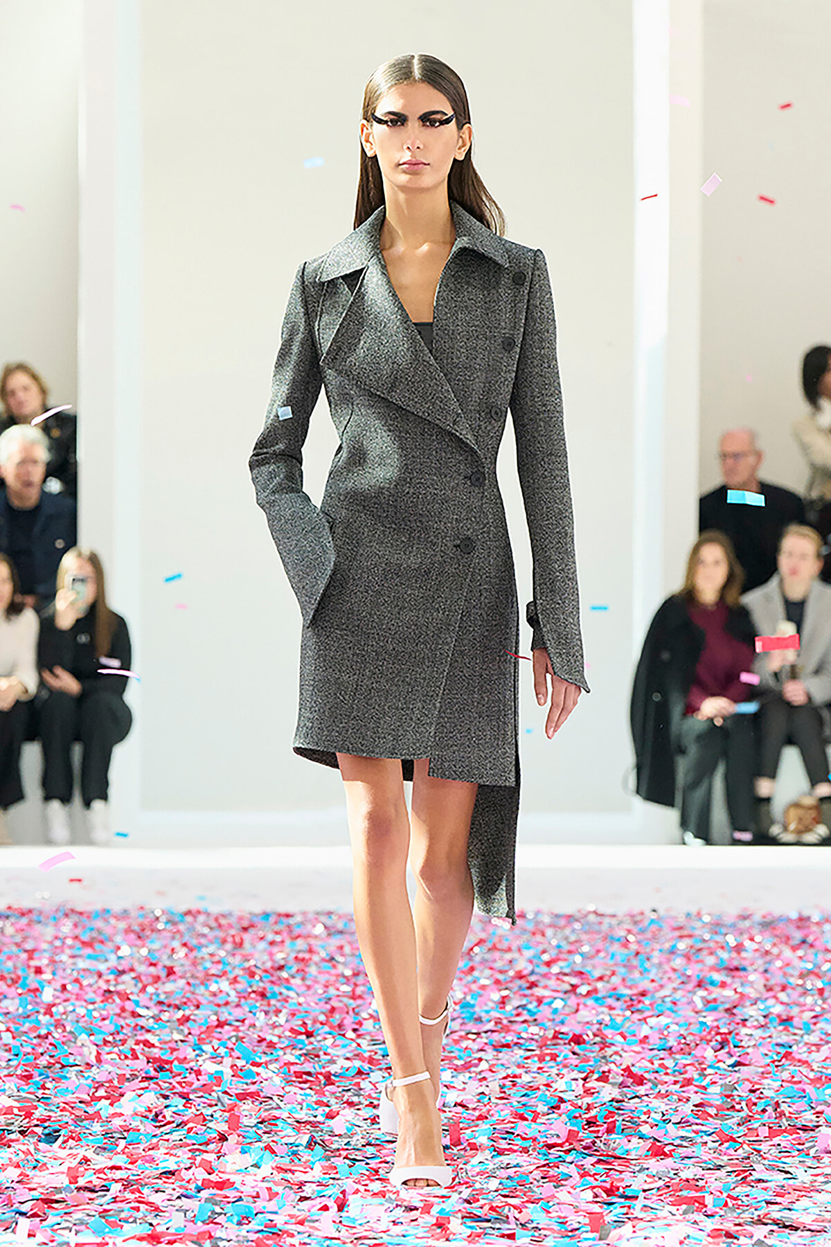 Model walks a fashion runway in a gray double-breasted coat; pink and blue confetti covers the floor.