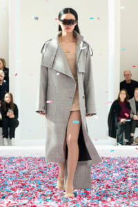 Model walking a runway in a light gray oversized coat over a nude dress, beige tights, and futuristic visor sunglasses as confetti falls around.