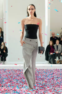 Model walks the runway in a black strapless top and gray wide‑leg pants, clutch in hand, as confetti falls around her.