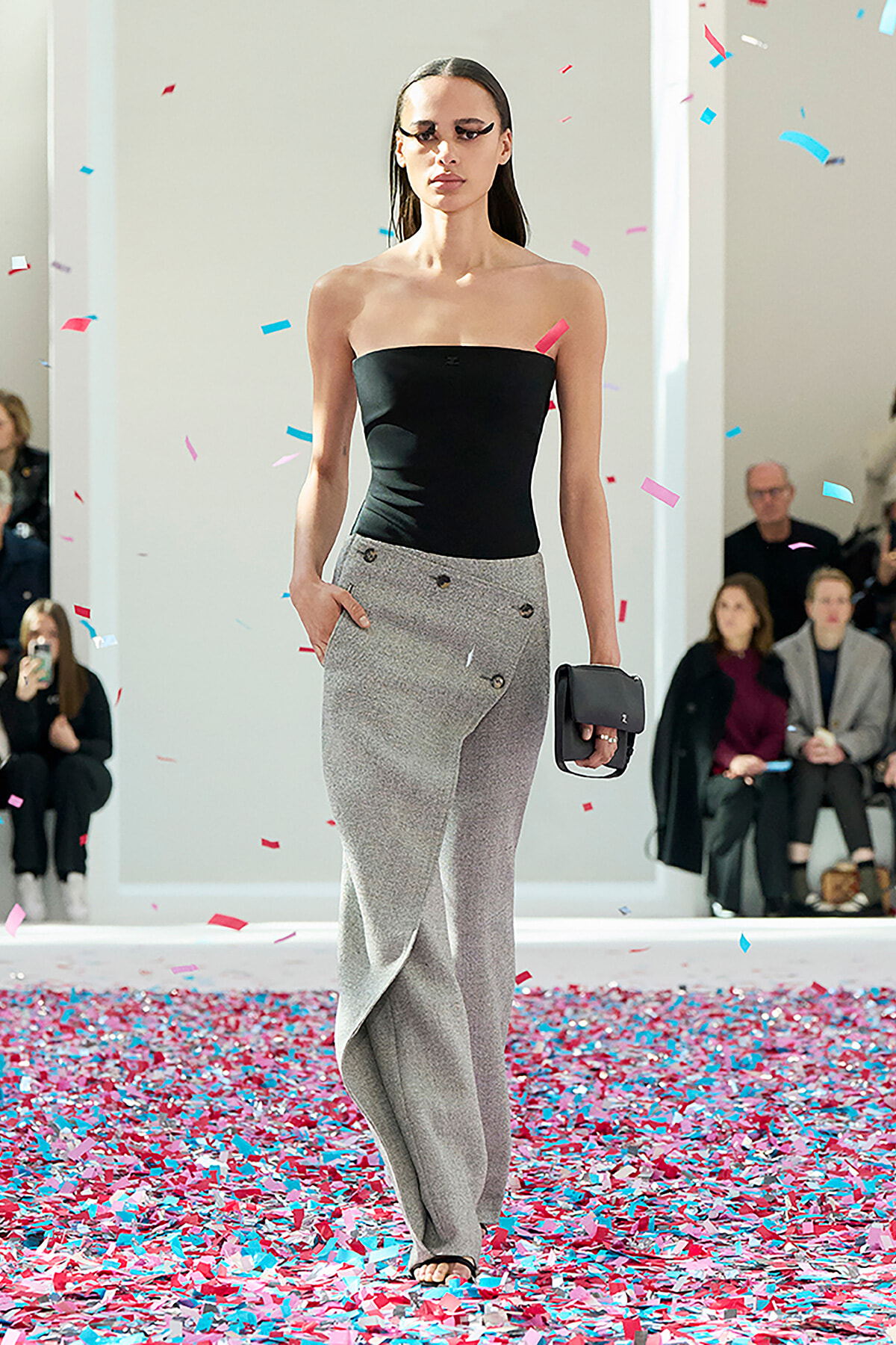 Model walks the runway in a black strapless top and gray wide‑leg pants, clutch in hand, as confetti falls around her.