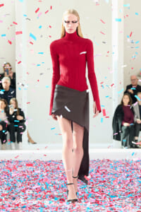 Model walks the runway in a red ribbed turtleneck and black asymmetrical leather skirt amid colorful confetti.