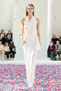 Model in a white halter jumpsuit walks a runway as pink and blue confetti falls around her; audience seated along the sides.