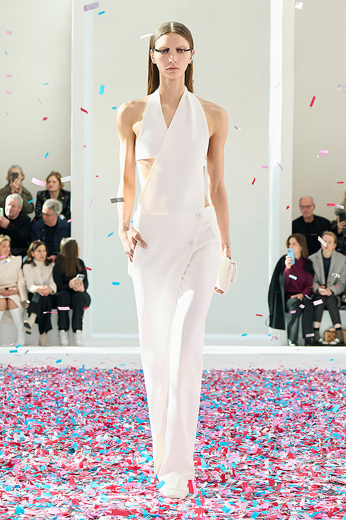 Model in a white halter jumpsuit walks a runway as pink and blue confetti falls around her; audience seated along the sides.