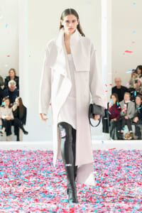 Model walks a runway in a pale pink oversized coat and black thigh-high boots, confetti falling around, audience in background.