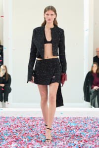 Model walking a runway in a speckled black cropped blazer and matching mini skirt with a black bra top, holding a red clutch amid confetti.
