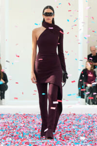 Model walking a runway in a burgundy one-shoulder knit dress with a matching scarf, wearing dark sunglasses as confetti falls around her.