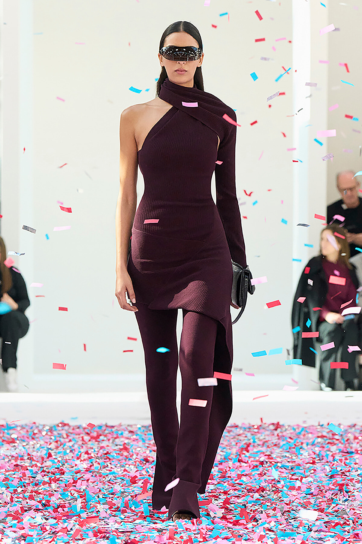 Model walking a runway in a burgundy one-shoulder knit dress with a matching scarf, wearing dark sunglasses as confetti falls around her.