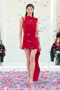 Model walking a fashion runway in a red asymmetrical dress amid blue and pink confetti