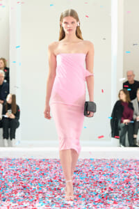 Model walks a pink one-shoulder dress down a confetti-covered runway, holding a small black clutch.