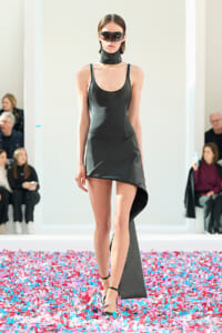 Model walking a black asymmetric mini dress on a fashion show runway, surrounded by colorful confetti and spectators in the background.
