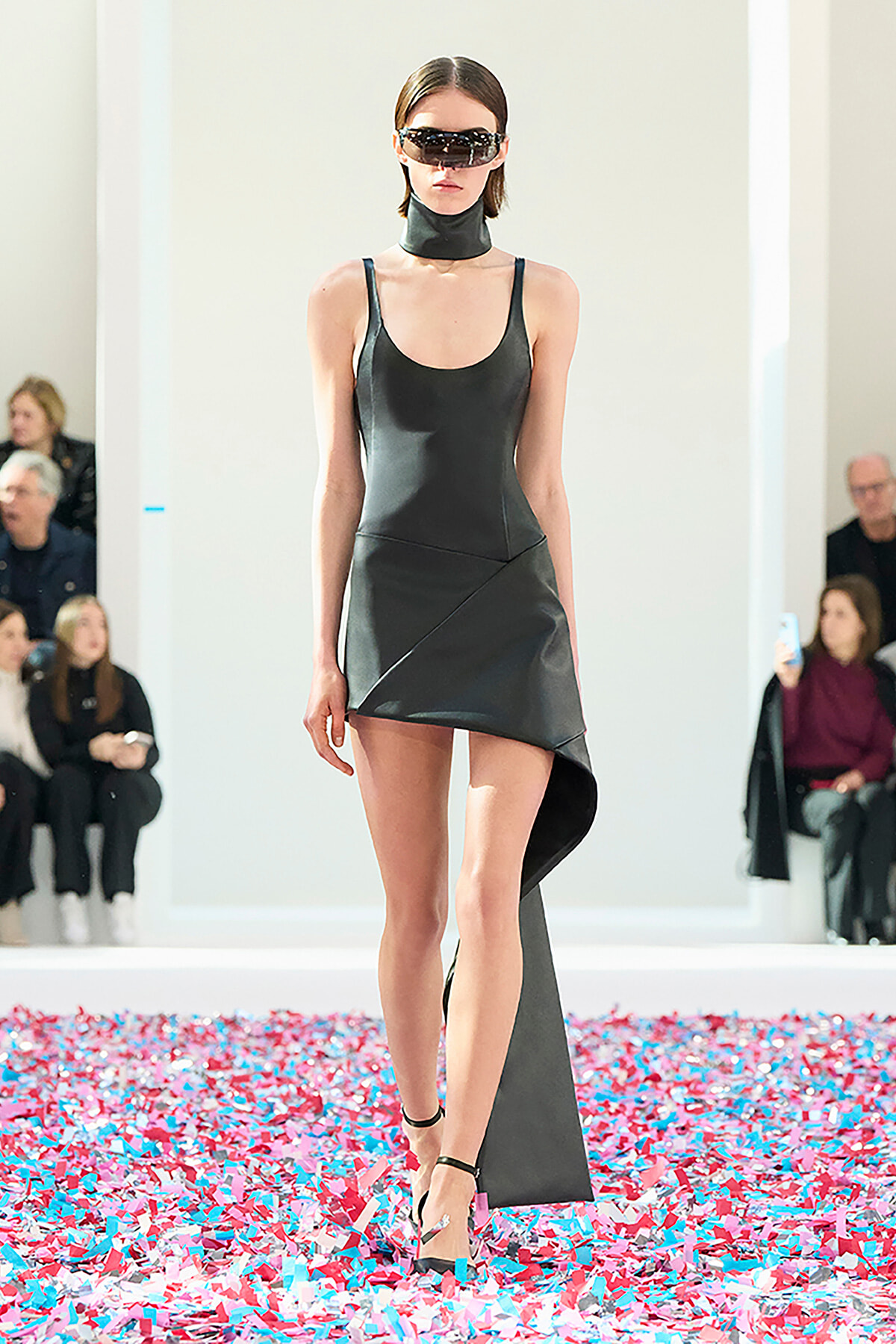 Model walking a black asymmetric mini dress on a fashion show runway, surrounded by colorful confetti and spectators in the background.