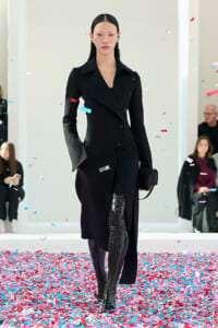 Model walks the runway in a black double‑breasted coat and patent thigh‑high boots amid colorful confetti fallyling the floor edges.