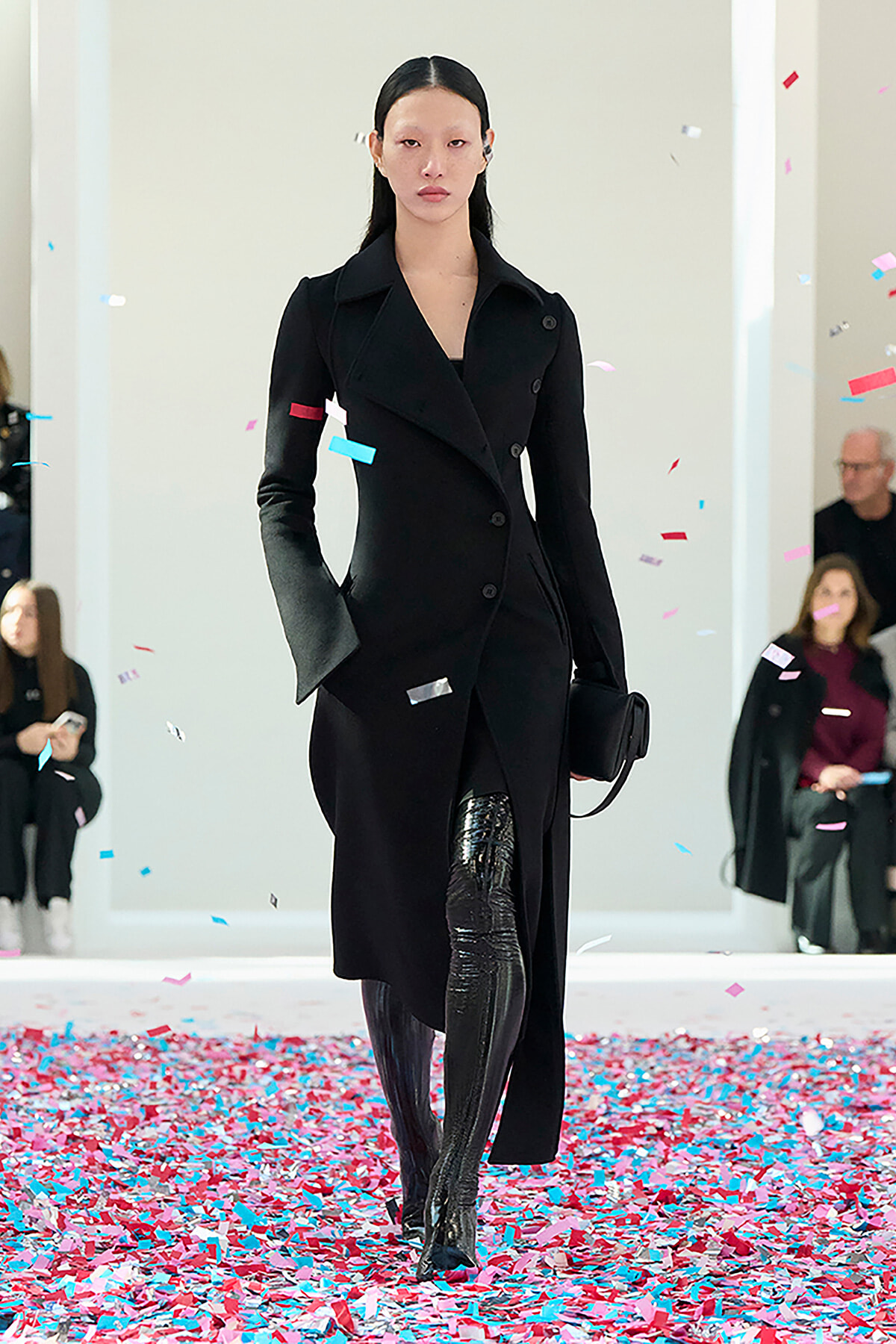 Model walks the runway in a black double‑breasted coat and patent thigh‑high boots amid colorful confetti fallyling the floor edges.