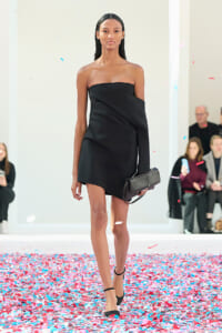 Model on a runway wearing a black off-shoulder mini dress with a matching black clutch, confetti on the floor nearby.