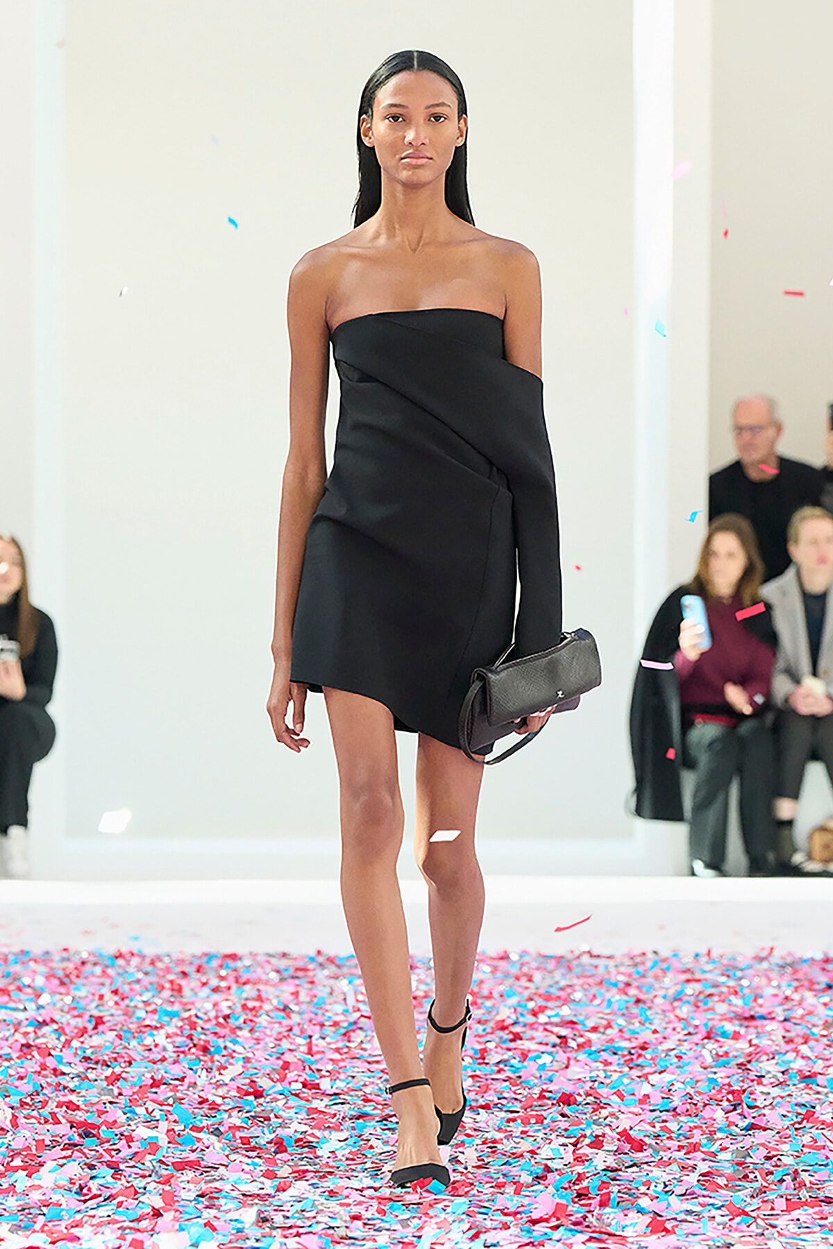 Model on a runway wearing a black off-shoulder mini dress with a matching black clutch, confetti on the floor nearby.