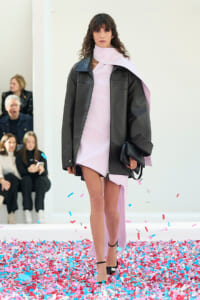 Model walking a fashion runway in a pale pink dress with a dark oversized jacket, holding a black clutch amid pink and blue confetti.