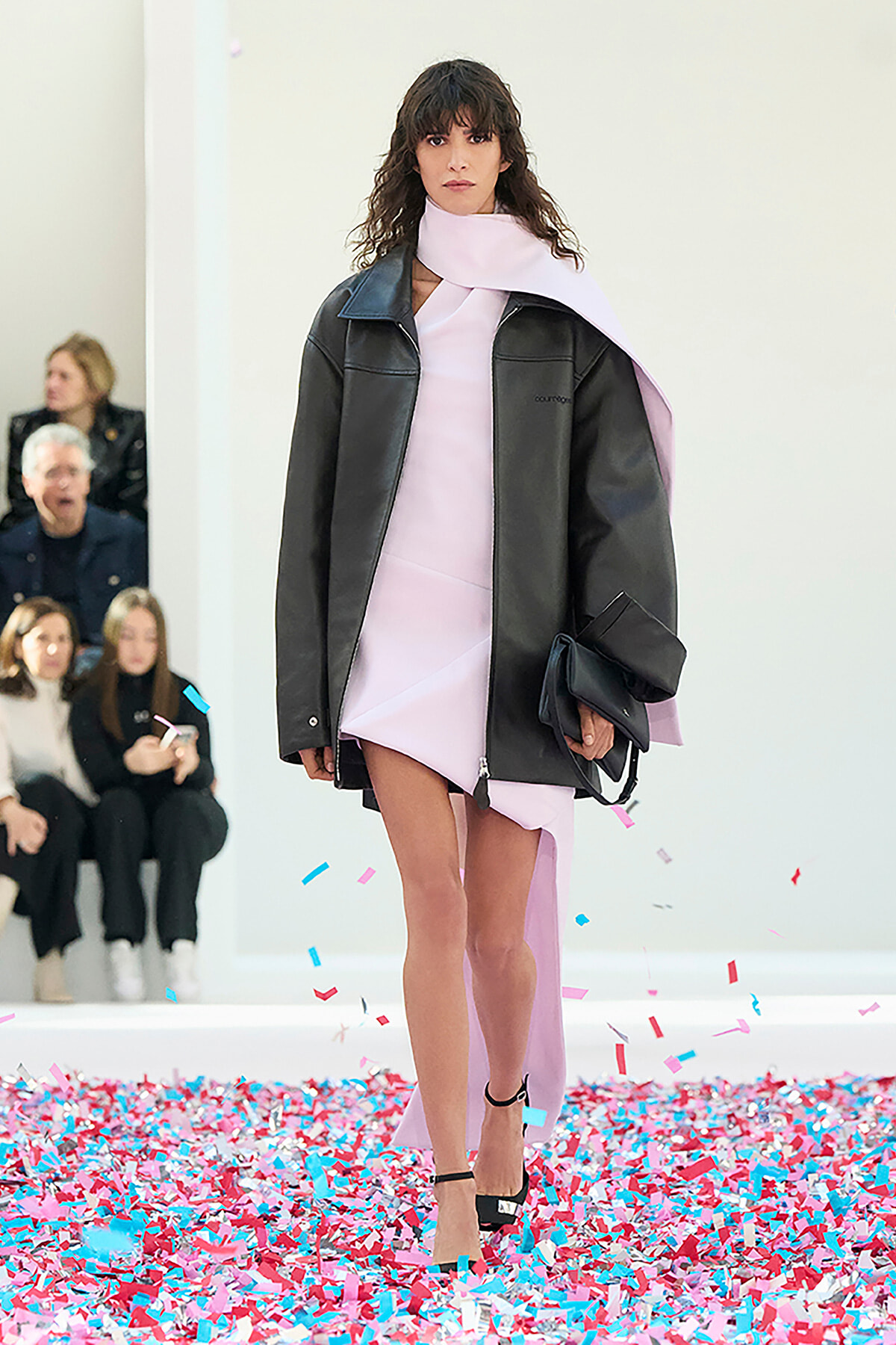 Model walking a fashion runway in a pale pink dress with a dark oversized jacket, holding a black clutch amid pink and blue confetti.