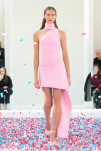 Model walking a runway in a light pink, one-shoulder halter dress with an asymmetric hem as pink and blue confetti falls around her.