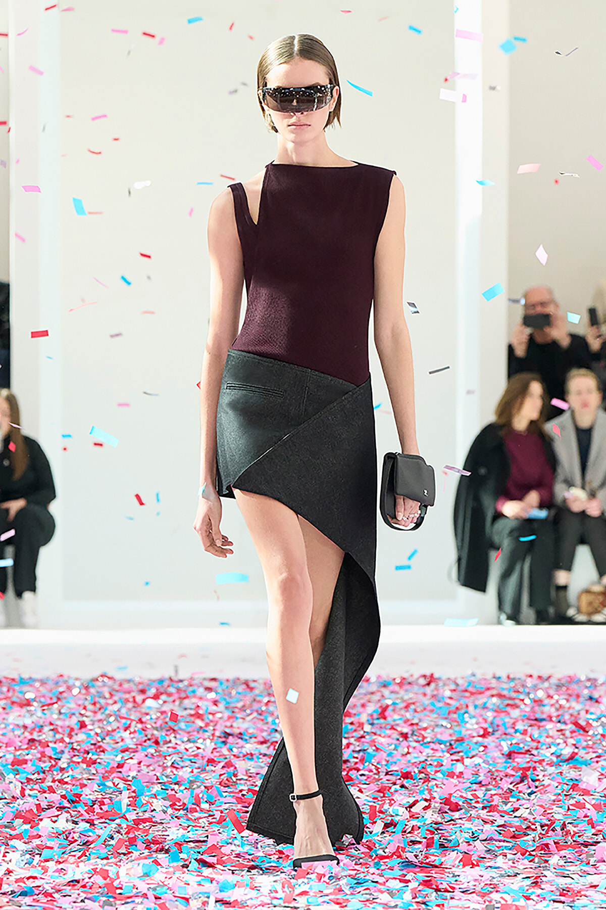 Model walking a runway in a burgundy sleeveless top and an asymmetric black skirt, wearing futuristic sunglasses and carrying a black clutch amid colorful confetti.