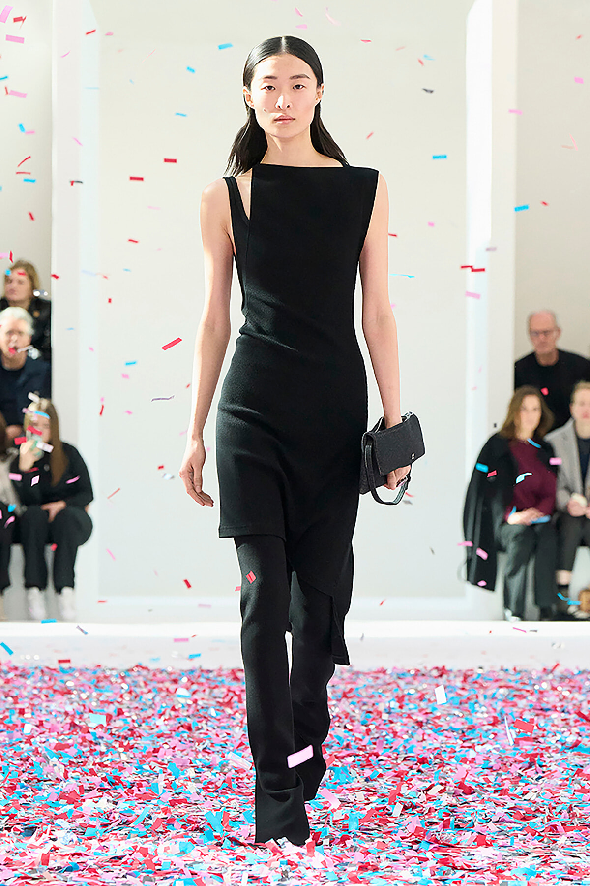 Model walks a runway in a sleeveless black dress, holding a small black clutch as pink and blue confetti falls around her.