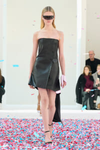 Model walks a runway in a black strapless leather wrap dress, futuristic sunglasses, and a pink clutch with colorful confetti on the floor.