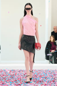 Model walking a runway in a pink one-shoulder top and black asymmetrical skirt, holding a red clutch amid confetti.