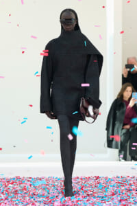 Model walking the runway in an oversized black cape-like top with high neck, black tights, and burgundy handbag, wearing futuristic sunglasses as colorful confetti falls.