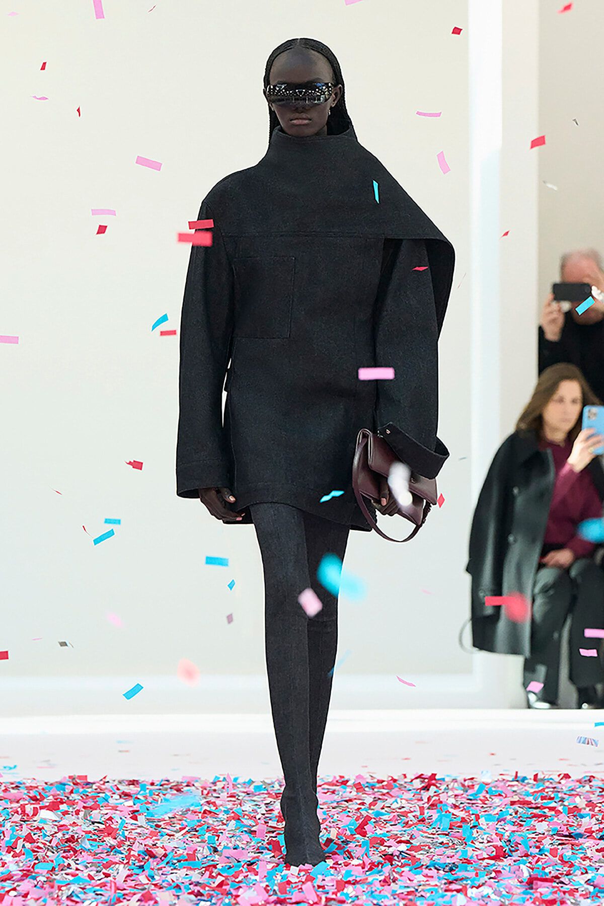 Model walking the runway in an oversized black cape-like top with high neck, black tights, and burgundy handbag, wearing futuristic sunglasses as colorful confetti falls.