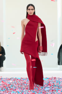 Model on a runway wearing a bold red, one-shoulder dress with a long scarf and matching tights, amid colorful confetti on the floor.