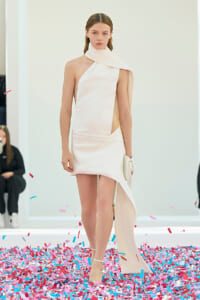 Model walks a runway in a minimalist white dress with a draped scarf detail and sandals, surrounded by pink and blue confetti on the floor.