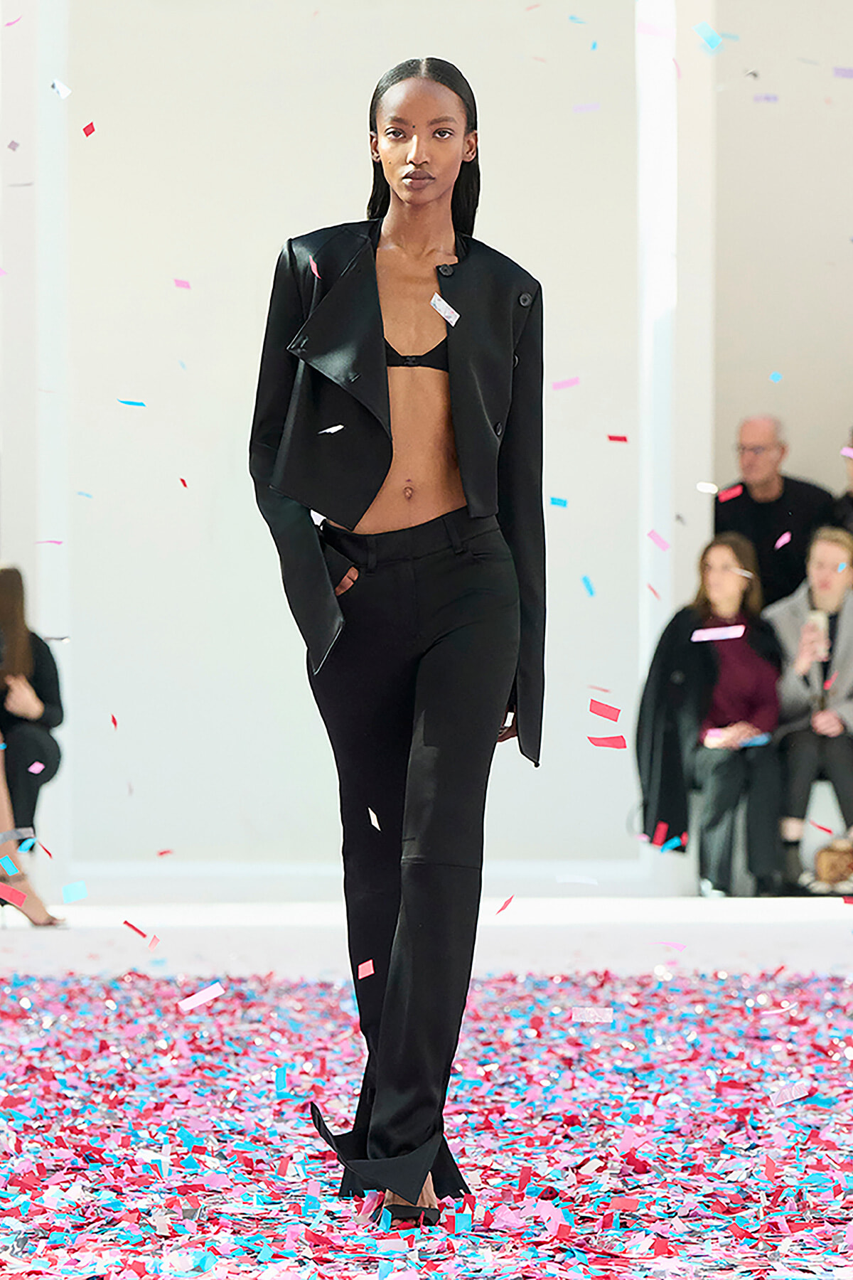Model walks the runway in a black cropped blazer and matching pants amid falling confetti.